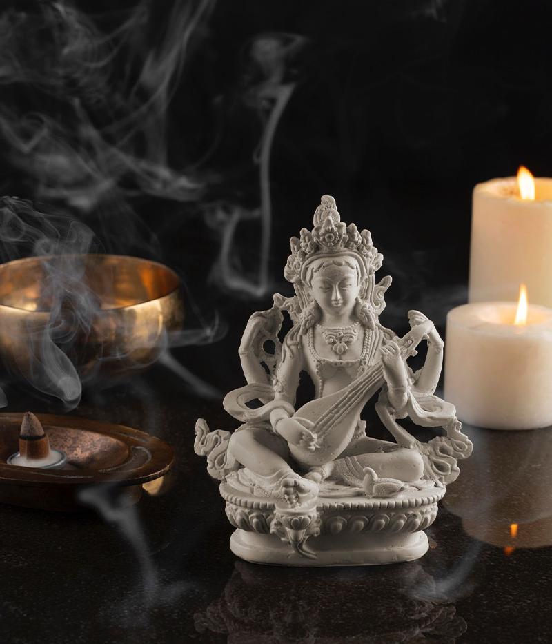 Statue of a deity with incense, candles, and a singing bowl on a reflective surface.
