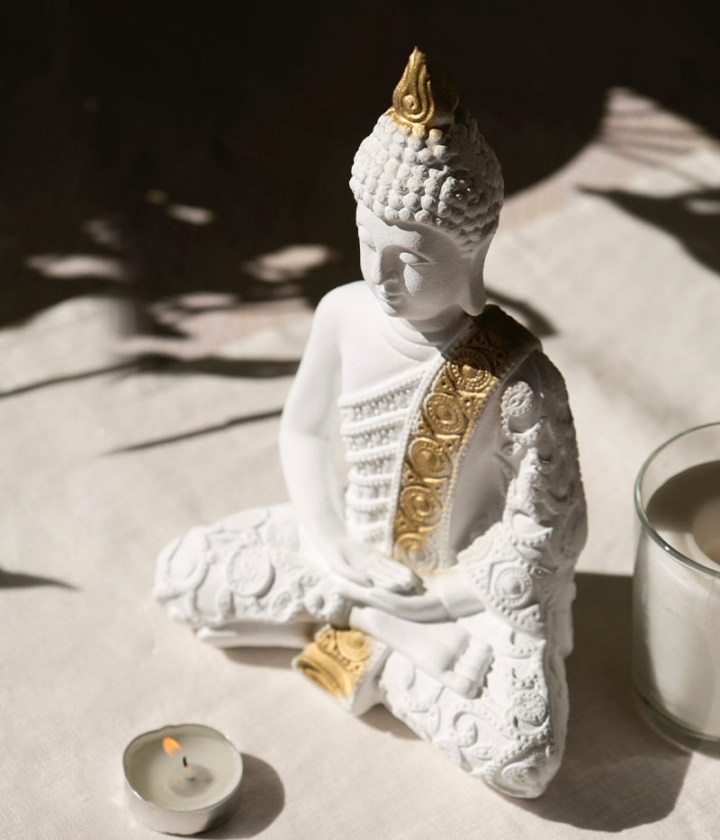 White Buddha statue with gold accents on a light surface with a candle and glass.